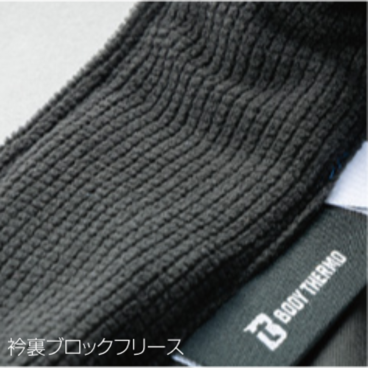 クロダルマ 54387 防風中綿ストレッチジャケットの衿裏ブロックフリース拡大画像。ブラックの凹凸あるフリース素材を衿裏に採用し、首元の保温性を高めた秋冬用防寒作業着。建設現場・物流倉庫・配送業・屋外作業など寒い季節の仕事着におすすめ。